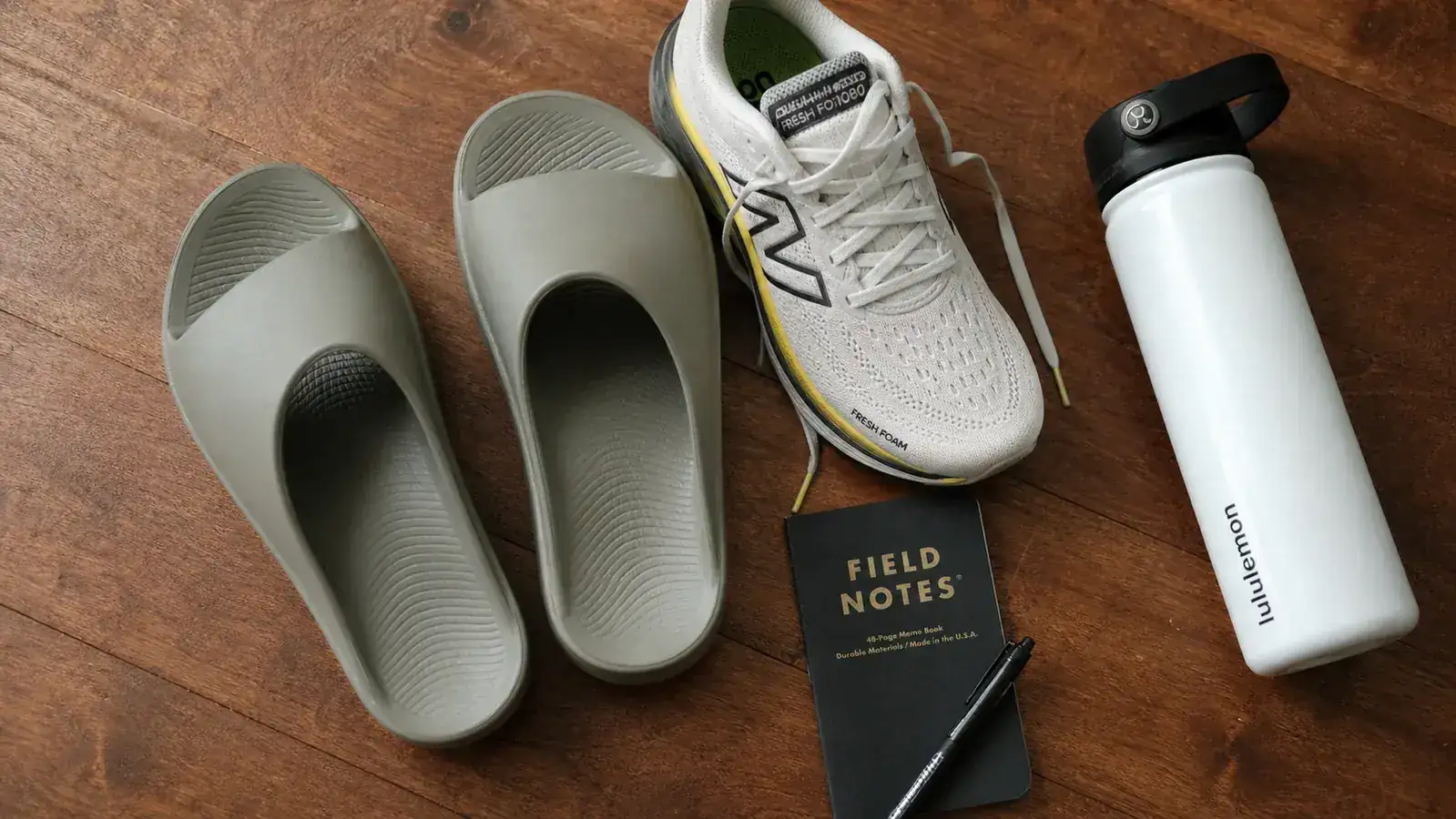 A pair of black OOFOS OOahh slides resting on a gym locker room bench next to a water bottle and a training bag